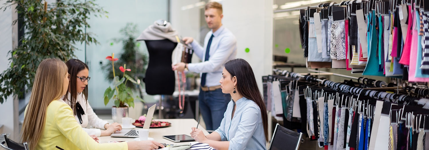 A workspace in a fashion studio where three people sit at a table reviewing materials, including a laptop, notebook, and fabric samples, while another person in the background adjusts a garment on a dress form.