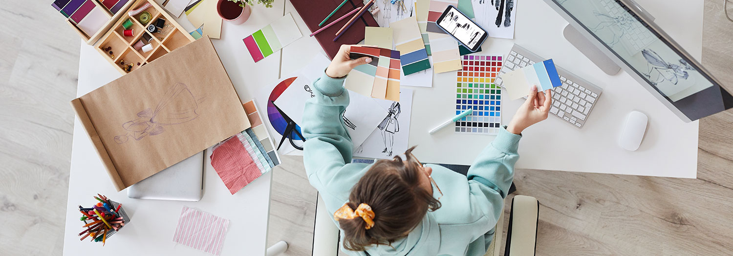 Overhead view of a person seated at a desk covered with creative materials, holding color swatches in both hands while comparing palettes. The workspace includes paint and fabric samples, colored pencils, sketches, a smartphone, keyboard, computer monitor and a paper bag with a fashion drawing.
