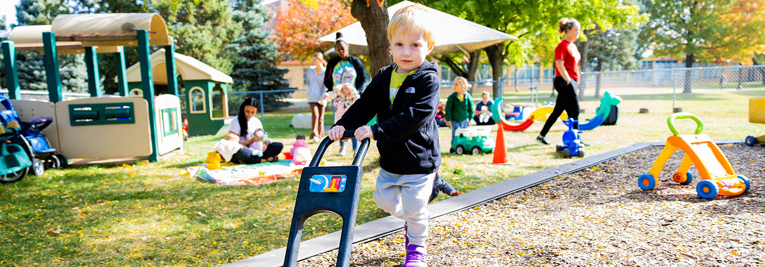 A toddler pushes a toy lawn mower at outdoor playground at  Child Development and Family Center. Other young children play with colorful ride‑on toys and walkers nearby, while adults supervise. 