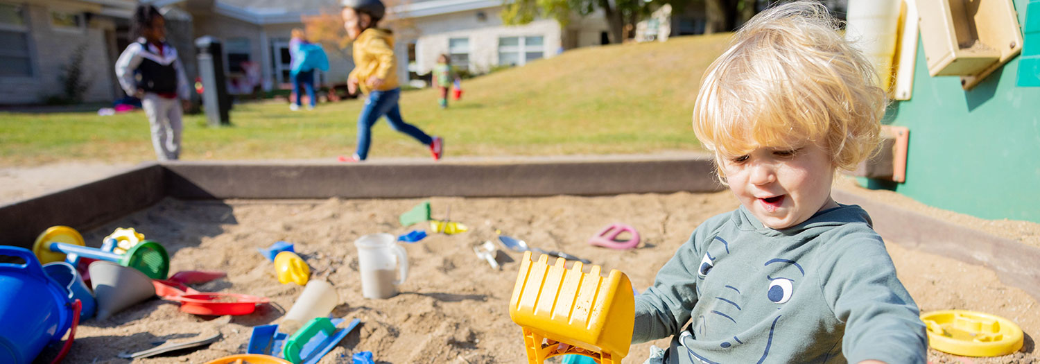 Toddler playing in a sandbox at Child Development and Family Center outdoor playground, with other children running and playing in the background