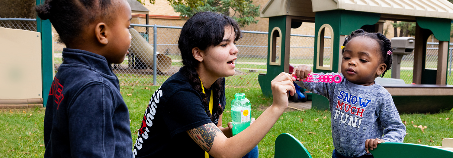 NIU student interacting with two toddlers on outdoor playground at Child Development and Family Center